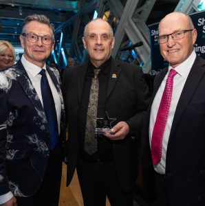 Three women standing outdoors. Two wear yellow T-shirts with a handprint logo, one holds a yellow balloon and wears glasses with a dark jacket. All are smiling at the camera.