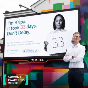 Richard Davidson standing in front of billboard featuring Kripa Shortt as part of Sarcoma Awareness Month 2024