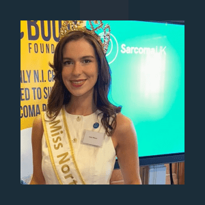 A young woman wearing a golden "Miss Northern Ireland" sash poses at what appears to be an awards ceremony or formal event. She has shoulder-length dark hair, is wearing a white sleeveless top with a name badge, and a decorative tiara or crown. Behind her is a turquoise display screen showing "Sarcoma UK" branding, and to the left is a yellow banner with partially visible text mentioning foundations and support services.