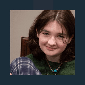A young person with shoulder-length brown hair smiles gently at the camera in what appears to be an indoor portrait setting. They're wearing a green jumper or cardigan over a blue and white checked shirt, with a distinctive beaded necklace featuring turquoise and other coloured stones.