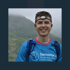 A young man wearing a bright blue Sarcoma UK charity t-shirt and headtorch stands smiling broadly outdoors. He has a blue rucksack and is positioned against a backdrop of misty green hills, suggesting he's on a hiking or outdoor challenge for the bone and soft tissue cancer charity.