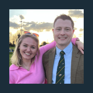 A woman in a pink jumper smiling with her arm around a man in a jacket and tie. They are outdoors with trees and the evening sky in the background.
