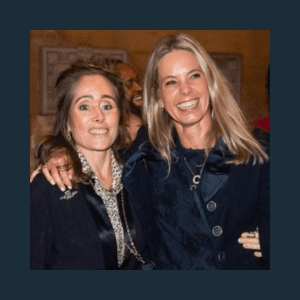 Two women smiling with their arms around each other. One wears a dark jacket with a patterned blouse, the other a dark coat. They are indoors with people in the background.
