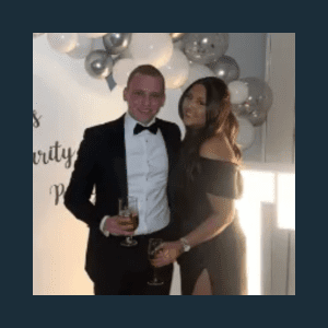 A man in a suit and bow tie stands beside a woman in a black evening dress. They are smiling and holding drinks, with balloons and a lit sign in the background.