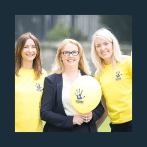 Three women standing outdoors. Two wear yellow T-shirts with a handprint logo, one holds a yellow balloon and wears glasses with a dark jacket. All are smiling at the camera.