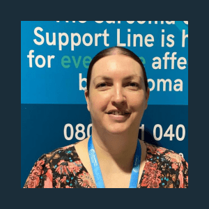 A woman smiling at the camera, wearing a patterned top and a lanyard. She is standing in front of a blue banner with white text that includes a helpline number.