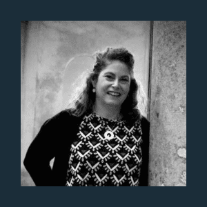 Black-and-white photo of a woman standing beside a stone wall. She is smiling, with curly hair, wearing a patterned dress and a dark cardigan.