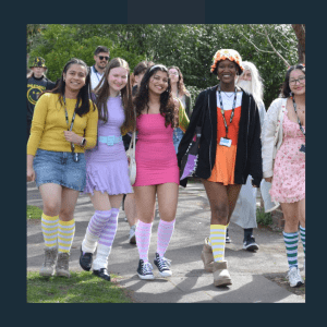 A group of young people wearing colourful themed costumes pose together outdoors at what appears to be a charity walk or festival event. They're all wearing matching striped knee-high socks in various colours (yellow, purple, pink), name badges, and bright clothing including yellow cardigans, purple and pink dresses, and orange outfits. Trees and other participants are visible in the background, with the group clearly taking part in some form of organised charity activity or fundraising event.