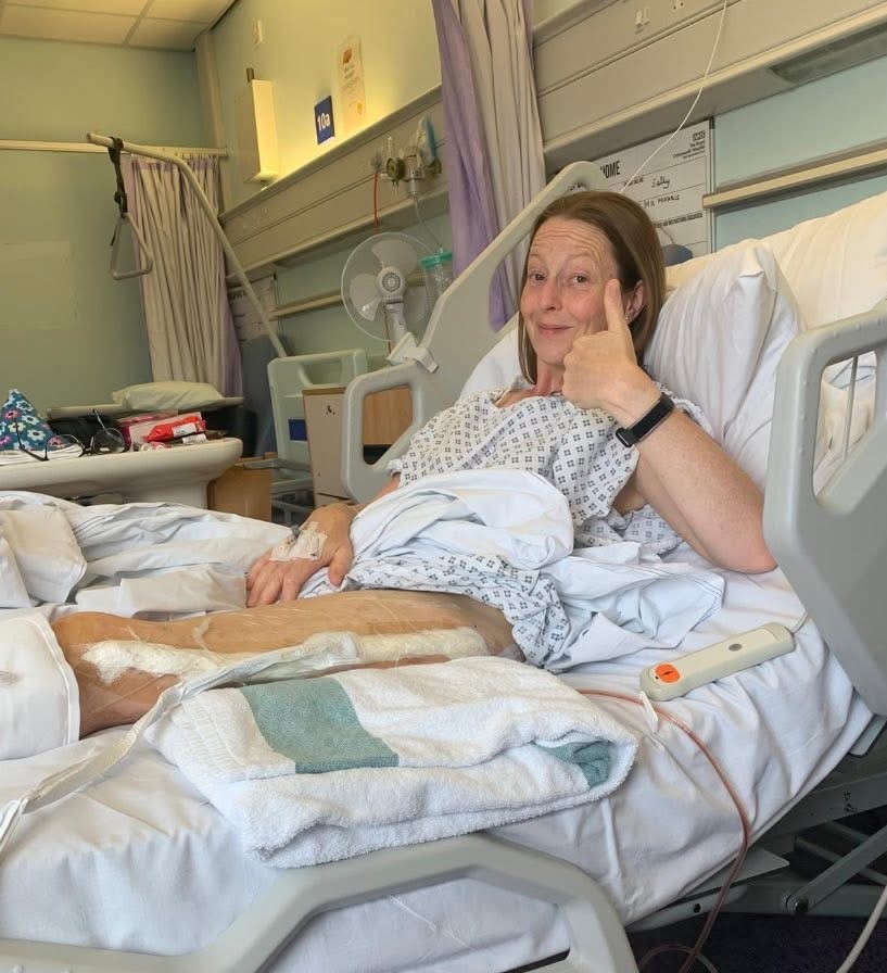 A woman is lying in a hospital bed giving a thumbs-up. She is wearing a hospital gown, with medical dressings on her leg and a cannula in her hand. The setting shows a typical hospital ward with medical equipment and curtains in the background.