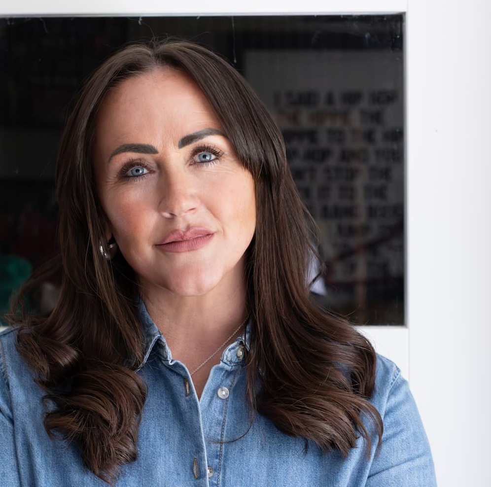 Anna Wright-Hicks looks towards the camera in a close-up portrait, wearing a light denim shirt against a softly blurred indoor background.