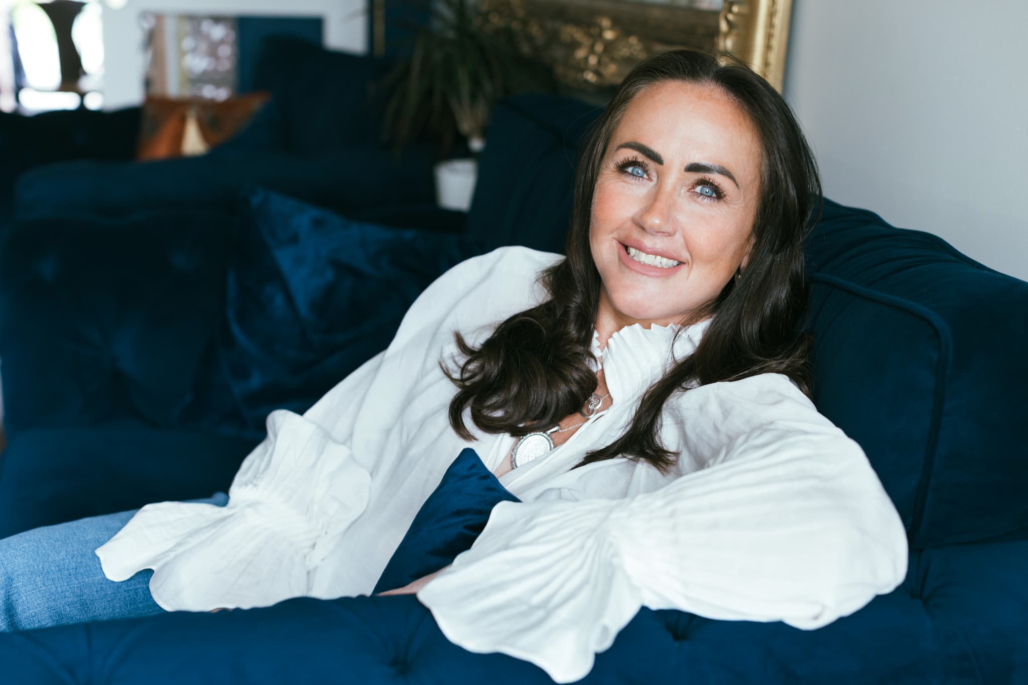 Anna Wright-Hicks smiles while reclining on a dark blue sofa, wearing a white blouse and jeans in a softly lit living room.