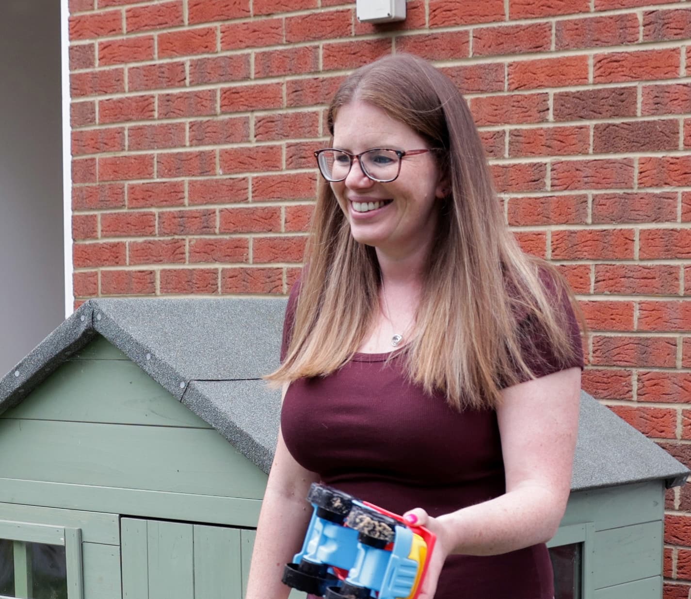 Woman with glasses smiling outside, holding a child’s toy truck beside a playhouse and brick wall.