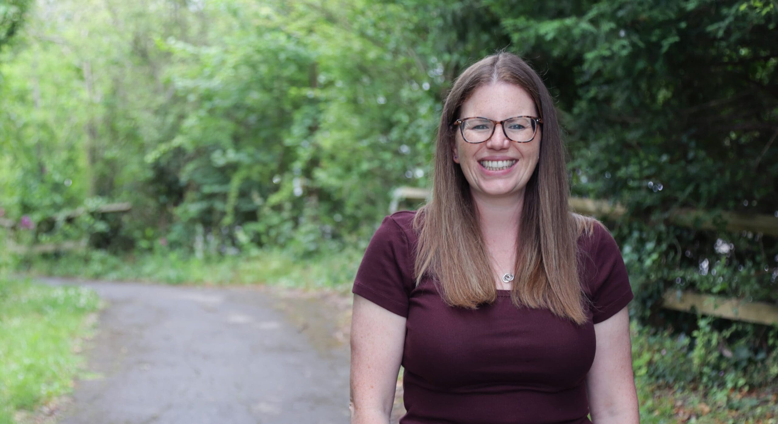 Woman with glasses smiling on a quiet path surrounded by trees and greenery.