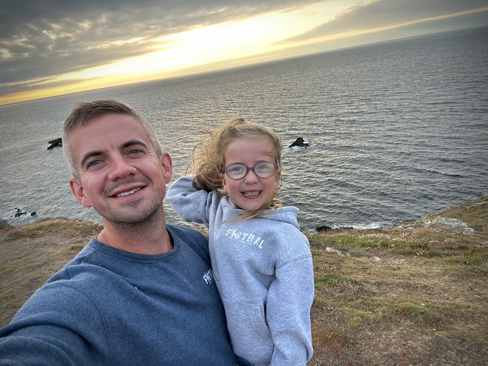 A man takes a selfie with a young child on a coastal cliff, with the sea and sunset behind them.