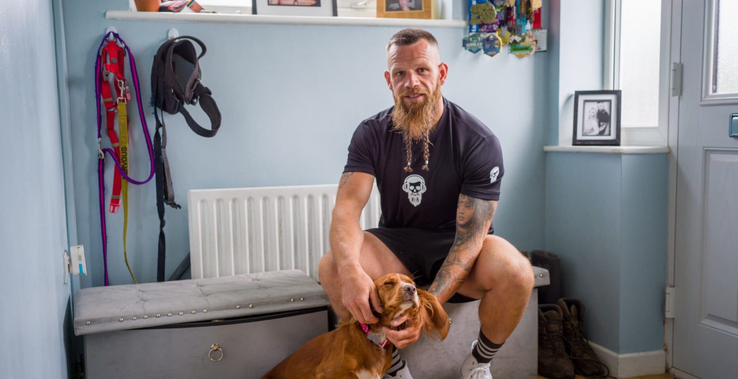 Robert Bell Johnson sits on a hallway bench at home, gently holding his small brown dog, with walking gear, family photos and medals visible around him.