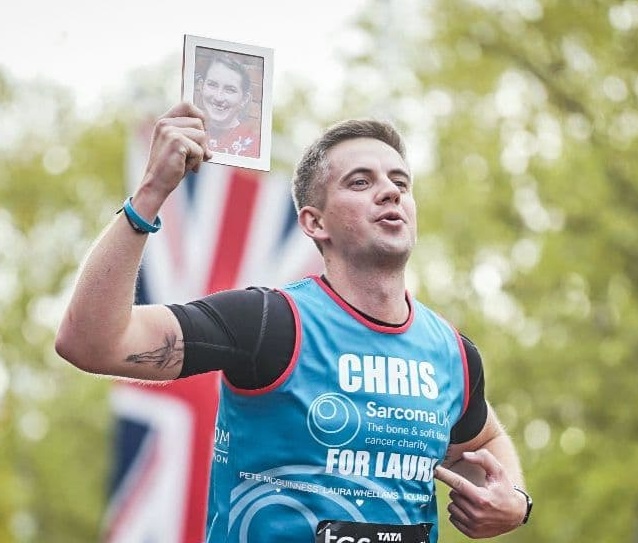 A medium close-up photograph of Chris Whellams running at a marathon event. He is wearing a blue Sarcoma UK charity running t-shirt printed with the words 