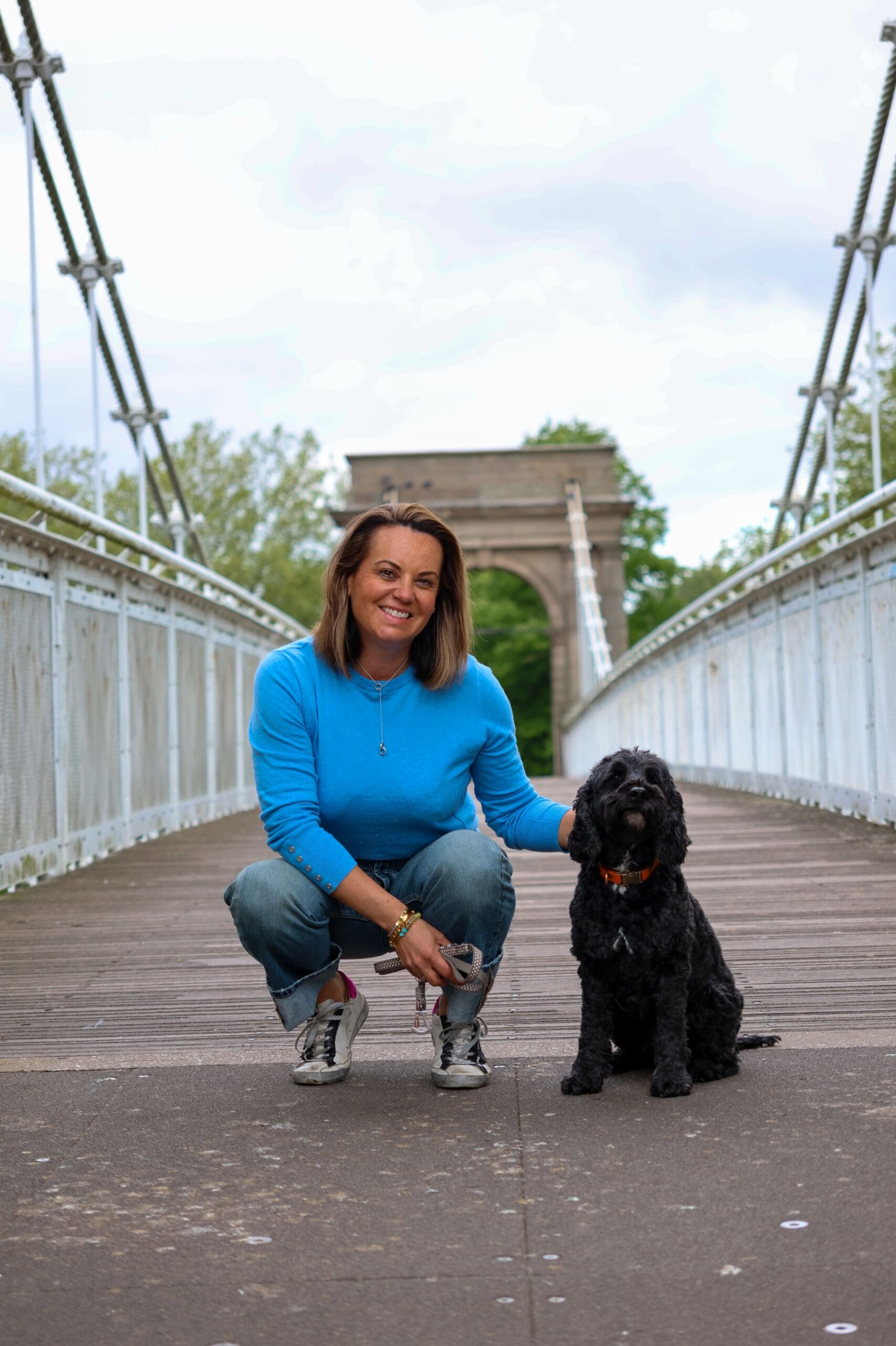A full-length outdoor photograph of Rachel Stevenson crouching on the wooden deck of a pedestrian suspension bridge. She is smiling at the camera, has shoulder-length brown hair, and is wearing a bright blue long-sleeved top, turned-up blue jeans, and grey trainers. Sitting upright beside her is her black cockapoo, Teddy, wearing an orange collar. Rachel's hand rests gently on the dog's back. The white metal railings of the bridge stretch back towards a grand stone archway structure with green trees visible in the distance.