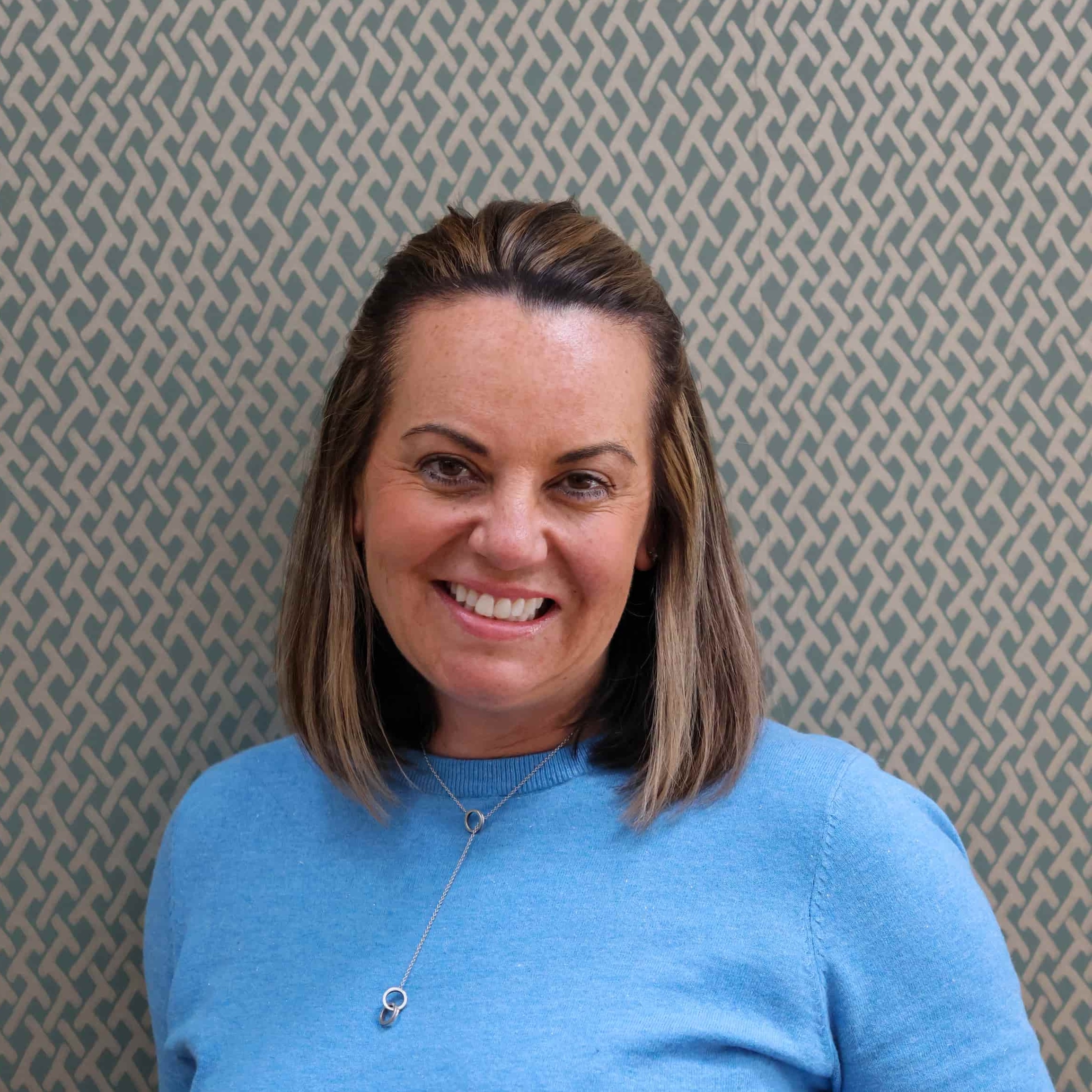 A head and shoulders portrait of Rachel Stevenson smiling directly at the camera. She has straight, shoulder-length brown hair and is wearing a vibrant blue crewneck jumper with a silver necklace, set against a background of green and grey geometric lattice-patterned wallpaper.