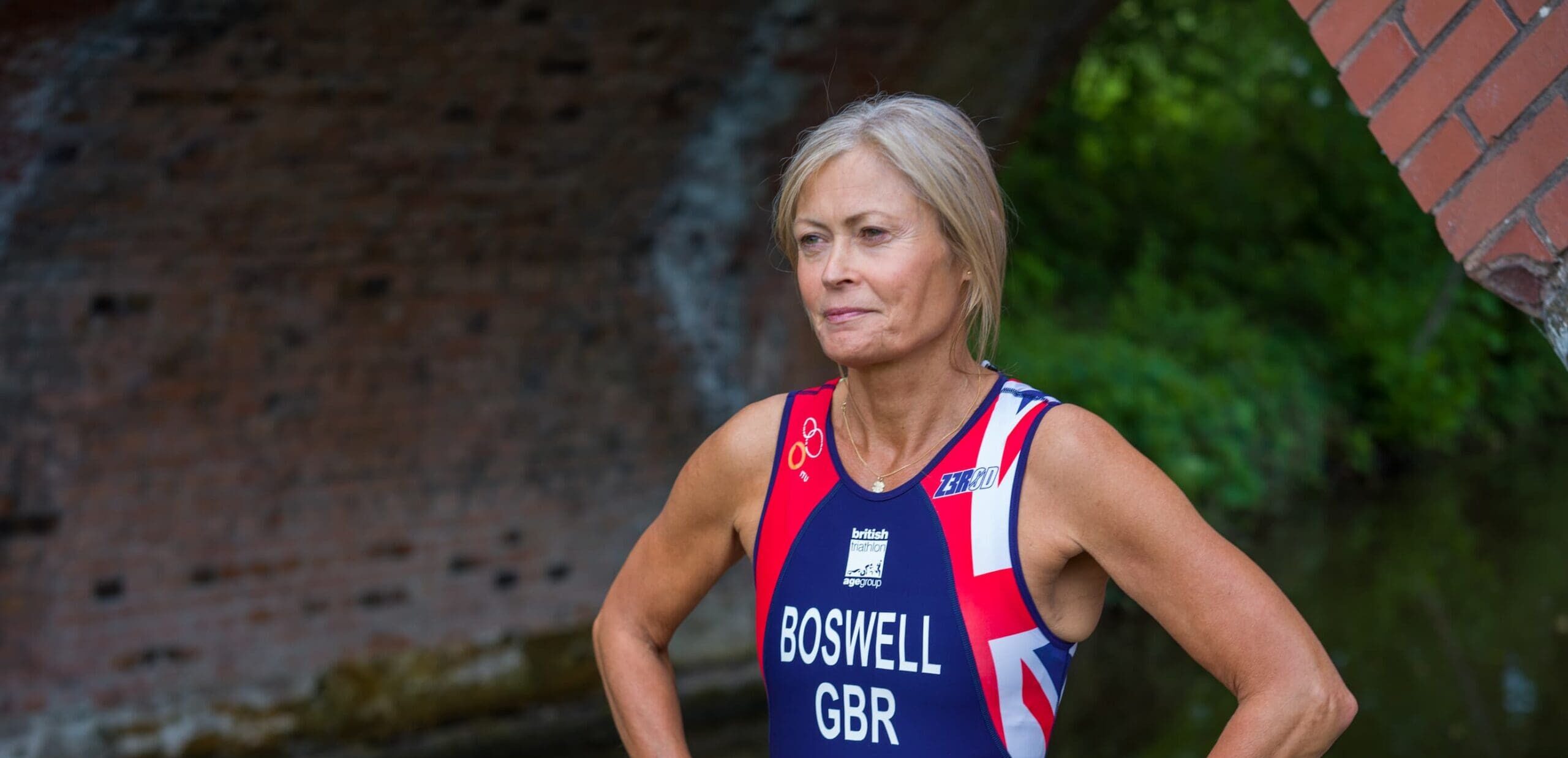 Samantha Boswell stands by a canal in a GB triathlon suit, hands on hips, looking ahead under a brick bridge.