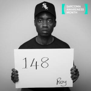 Black and white portrait of Roy Mugera looking directly at the camera while holding a white board displaying the number "148" and his name, "Roy". He is wearing a black baseball cap and dark top. The Sarcoma Awareness Month logo appears in the top right corner.