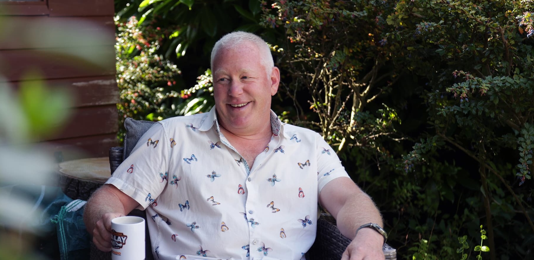 Man sitting in a garden chair, smiling and holding a mug, surrounded by plants and shrubs.