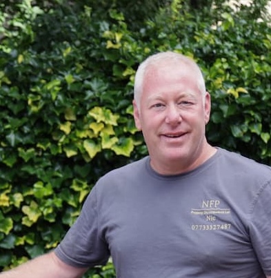 Man standing outdoors in front of leafy green hedges, wearing a grey T-shirt.