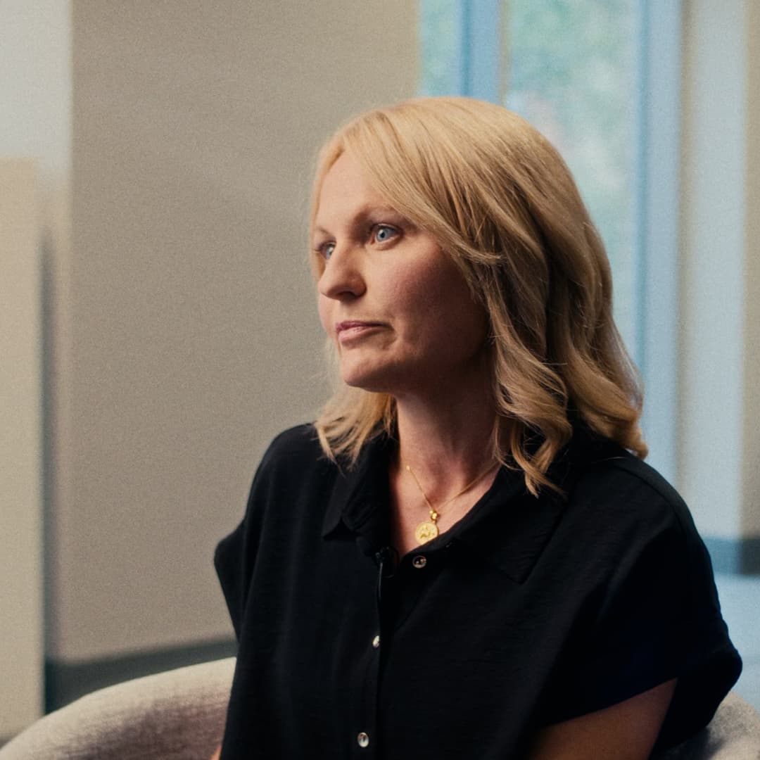 Portrait of Carly McDonald with shoulder-length blonde hair, wearing a black top and seated indoors, looking slightly to her right.
